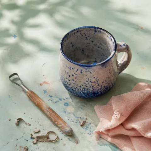 A speckled handmade mug on a mint pottery table beside a tool and pink cloth, with animated highlights.
